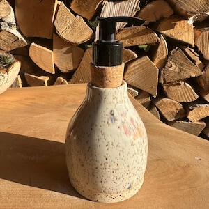 Könnte beinhalten: Ein gesprenkelter Seifenspender aus Keramik mit schwarzer Pumpe und Korkbasis. Der cremefarbene Spender hat eine bauchige Form und ist mit kleinen, farbenfrohen floralen Akzenten verziert. Der Spender steht auf einer Holzoberfläche.