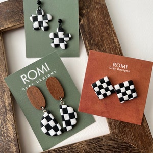 Puede incluir: Tres pares de pendientes con estampado de tablero de ajedrez en tarjetas de exhibición. Las tarjetas están etiquetadas con "ROMI Clay Designs". Los pendientes están hechos de arcilla polimérica y presentan patrones de tablero de ajedrez en blanco y negro en varias formas, incluyendo flores, cuadrados y semicírculos.