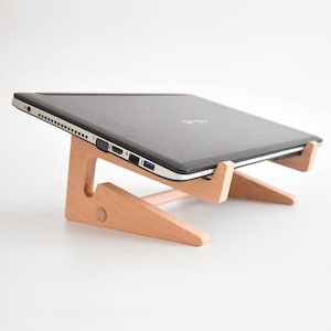 May include: A wooden laptop stand with a laptop resting on top. The stand is made of light wood and has a simple design. The laptop is black and has a silver trim.