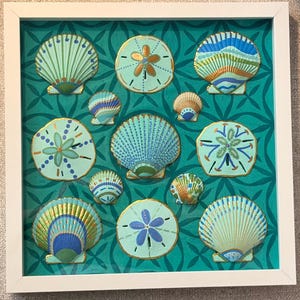 May include: A square white frame containing a collection of hand-painted seashells and sand dollars arranged on a teal background with a repeating geometric pattern. The shells feature intricate blue, green, and gold painted details.