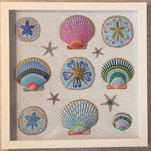 May include: A square white frame containing a collection of hand-painted seashells and sand dollars arranged on a light blue and white wavy patterned background. The shells feature vibrant, multi-colored designs with gold-painted edges.