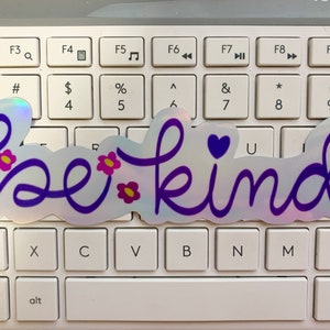 Puede incluir: Una pegatina hologr&aacute;fica con las palabras "be kind" en escritura morada. La pegatina presenta peque&ntilde;os acentos de flores rosas y amarillas y un coraz&oacute;n morado. La pegatina est&aacute; colocada sobre un teclado blanco.