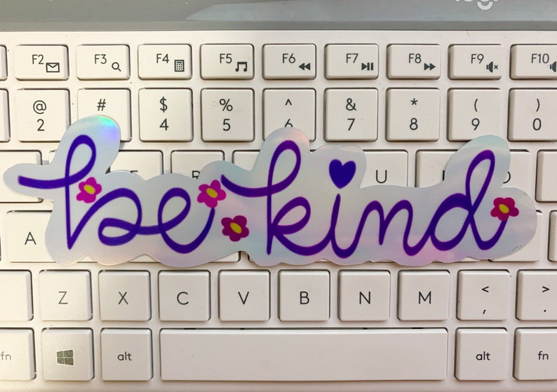 Puede incluir: Una pegatina hologr&aacute;fica con las palabras "be kind" en escritura morada. La pegatina presenta peque&ntilde;os acentos de flores rosas y amarillas y un coraz&oacute;n morado. La pegatina est&aacute; colocada sobre un teclado blanco.