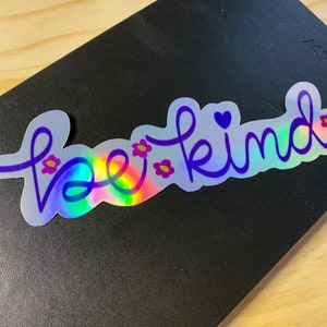 Puede incluir: Pegatina hologr&aacute;fica con las palabras "be kind" en escritura morada. La pegatina tiene un brillo de arco&iacute;ris y est&aacute; decorada con peque&ntilde;as flores rosas y un coraz&oacute;n morado. La pegatina est&aacute; sobre una superficie negra.