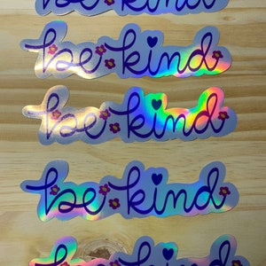 Puede incluir: Cinco pegatinas hologr&aacute;ficas con la frase "be kind" en cursiva. Las pegatinas tienen un contorno azul, peque&ntilde;as flores rosas y un coraz&oacute;n. El fondo es de madera de color marr&oacute;n claro.
