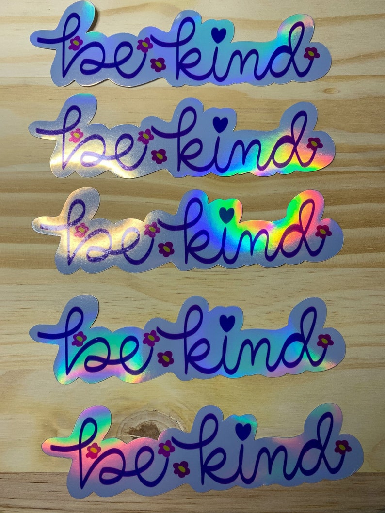 Puede incluir: Cinco pegatinas hologr&aacute;ficas con la frase "be kind" en cursiva. Las pegatinas tienen un contorno azul, peque&ntilde;as flores rosas y un coraz&oacute;n. El fondo es de madera de color marr&oacute;n claro.