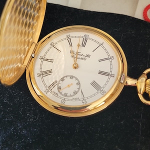Pode incluir: Um relógio de bolso dourado com mostrador branco e algarismos romanos pretos. O relógio tem um pequeno mostrador de segundos e o texto "E. Dissot & Fils Depuis 1853". O relógio está aberto, revelando o design intrincado da caixa de ouro.