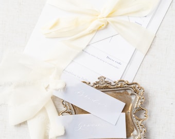 Può includere: Due biglietti bianchi con le parole "Bride" e "Groom" scritte in calligrafia sono esposti su un piccolo vassoio dorato decorato. Il vassoio è su una superficie bianca testurizzata. Un nastro bianco è legato con un fiocco sopra una pila di buste bianche.