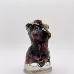 May include: A decorative resin torso sculpture. The sculpture is filled with a mix of dark pink, green, and black stones. The base is clear, and the torso is a glossy finish. The sculpture is a unique piece of art.