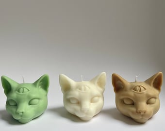 Puede incluir: Tres velas con forma de cabeza de gato, una verde, una blanca y una marrón. Cada vela tiene un tercer ojo en el centro de la frente.