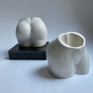 May include: Two white ceramic sculptures shaped like human buttocks. The sculptures are on a black square base.