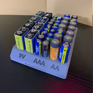 Könnte beinhalten: Ein grauer, 3D-gedruckter Batterie-Organizer, der verschiedene Batterien enthält, darunter 9V-, AAA- und AA-Größen. Der Organizer hat Etiketten für jeden Batterietyp. Batterien sind in verschiedenen Farben, darunter Gelb, Blau und Orange.