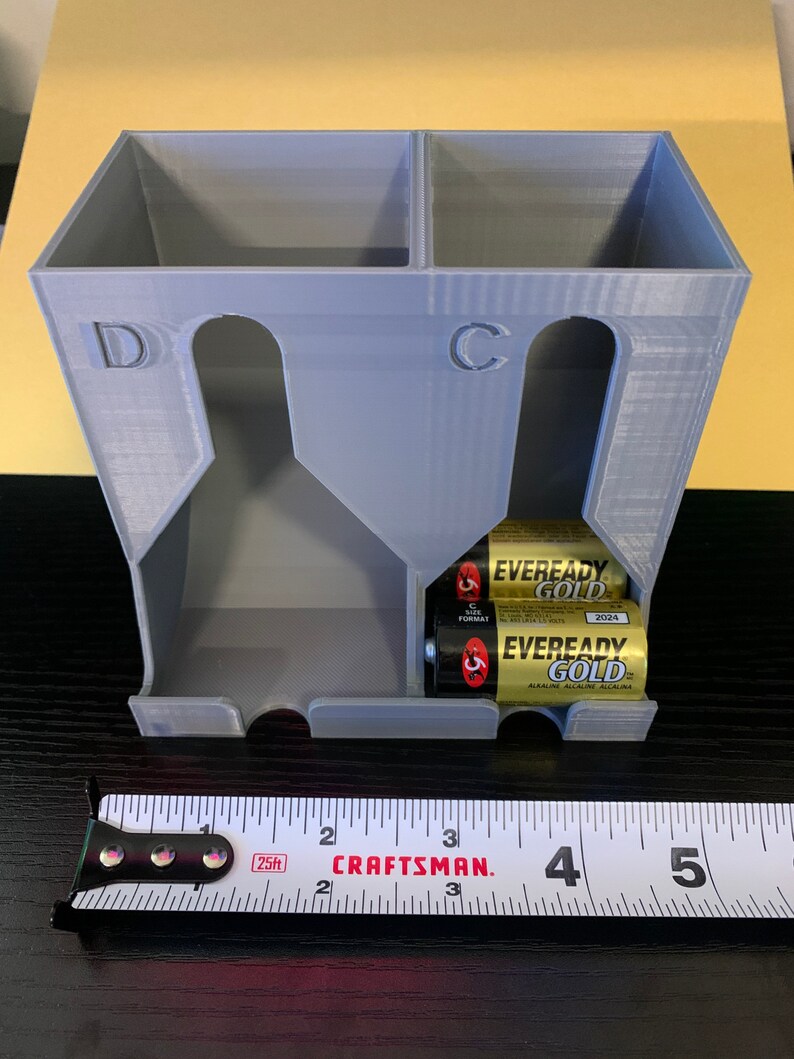 May include: A gray plastic battery organizer with two compartments labeled "D" and "C". The organizer is holding two sets of batteries, one set of "C" size batteries and one set of "D" size batteries. The batteries are Eveready Gold brand.
