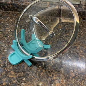 May include: Teal plastic lid holder for a glass pot lid. The lid holder has a curved design to hold the lid securely.