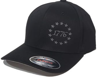 May include: Black baseball cap with a gray embroidered circle of stars and the text "1776". The cap has a Flexfit label on the back.