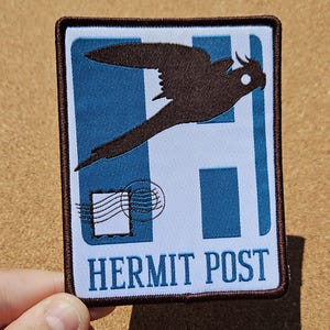 May include: Embroidered patch with a brown bird in flight over a blue and white background. The patch features a postage stamp design and the words "HERMIT POST" in blue. The patch is rectangular with a brown border.