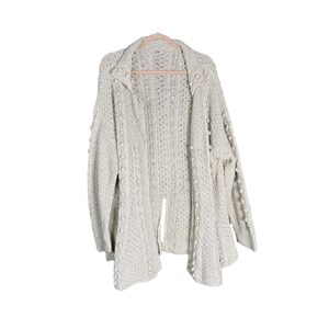 May include: A cream-colored, open-front cardigan sweater with a textured knit design. The sweater features a collar and is adorned with small, round bobbles along the sleeves and edges. It hangs on a wooden hanger against a white background.