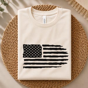 May include: A cream-colored crew neck t-shirt featuring a black graphic print of the American flag. The flag design has a distressed, brushstroke appearance with a rectangular field of stars on the left and horizontal stripes on the right.