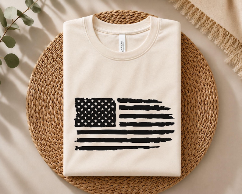 May include: A cream-colored crew neck t-shirt featuring a black graphic print of the American flag. The flag design has a distressed, brushstroke appearance with a rectangular field of stars on the left and horizontal stripes on the right.