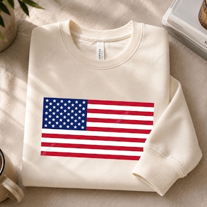 May include: A cream-colored crewneck sweatshirt featuring a graphic of the United States flag centered on the chest. The flag displays the traditional blue field with white stars and alternating red and white horizontal stripes. The garment has long sleeves.