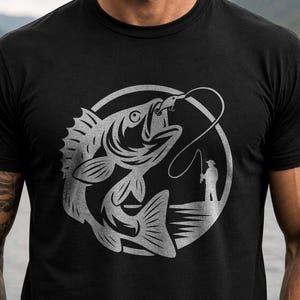 May include: A black short-sleeved t-shirt featuring a large, circular silver graphic on the chest. The design depicts a detailed fish hooked on a line, with a small silhouette of a person standing on a shoreline holding a fishing rod nearby.