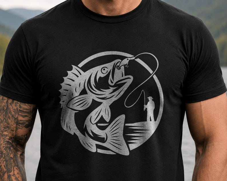 May include: A black short-sleeved t-shirt featuring a large, circular silver graphic on the chest. The design depicts a detailed fish hooked on a line, with a small silhouette of a person standing on a shoreline holding a fishing rod nearby.