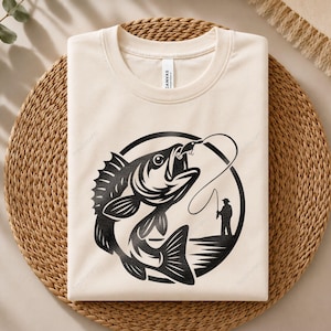 May include: A cream-colored crew neck t-shirt featuring a black graphic print of a large fish jumping out of the water. The design includes a silhouette of a person fishing on a shoreline, all enclosed within a circular border on the front chest.