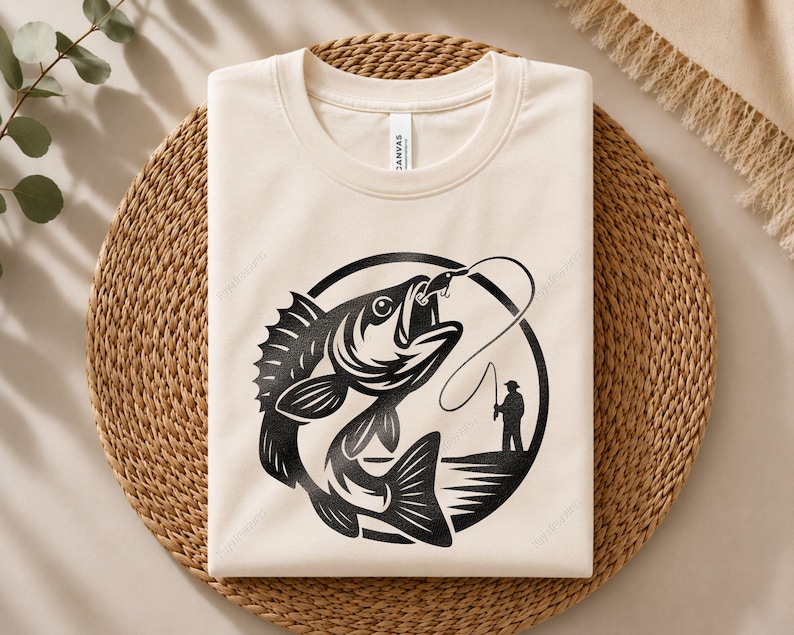 May include: A cream-colored crew neck t-shirt featuring a black graphic print of a large fish jumping out of the water. The design includes a silhouette of a person fishing on a shoreline, all enclosed within a circular border on the front chest.