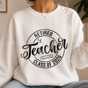 May include: A white long-sleeve sweatshirt featuring a black circular graphic on the chest. The text reads "RETIRED Teacher CLASS OF 2026" in a mix of block and script fonts, with a small pencil illustration centered inside the circle design.