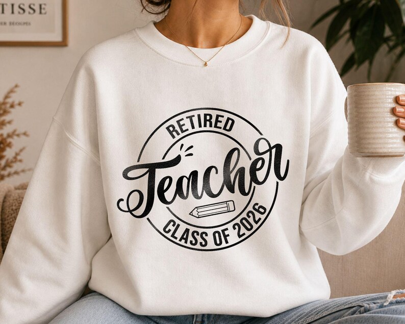 May include: A white long-sleeve sweatshirt featuring a black circular graphic on the chest. The text reads "RETIRED Teacher CLASS OF 2026" in a mix of block and script fonts, with a small pencil illustration centered inside the circle design.