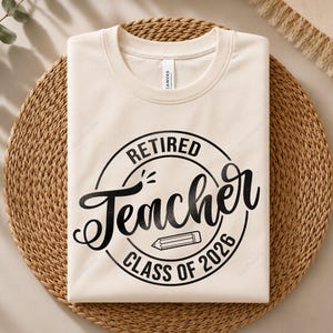 May include: A cream-colored short-sleeve t-shirt featuring a black circular graphic with the text "RETIRED Teacher CLASS OF 2026" and a small pencil icon in the center. The shirt has a crew neckline and is folded neatly for display.
