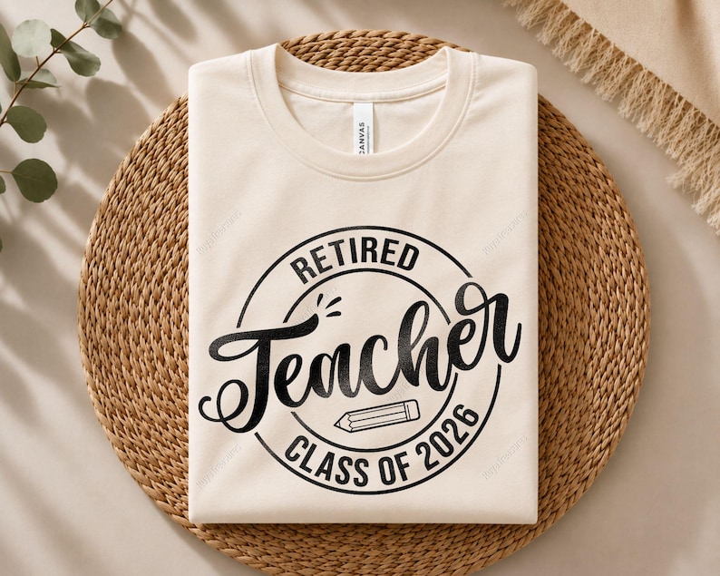 May include: A cream-colored short-sleeve t-shirt featuring a black circular graphic with the text "RETIRED Teacher CLASS OF 2026" and a small pencil icon in the center. The shirt has a crew neckline and is folded neatly for display.