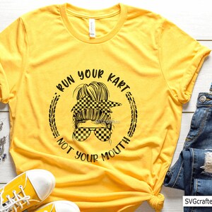 May include: Yellow t-shirt with a black and white checkered design of a woman with a messy bun and sunglasses. The text "Run Your Kart Not Your Mouth" is written around the design.