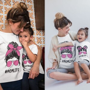 Peut inclure: T-shirts blancs avec des graphiques assortis. Les chemises pr&eacute;sentent le visage d'une femme avec un bandeau rose et des lunettes de soleil, et le texte "#MOM LIFE". Le t-shirt d'un enfant dit "#KID LIFE".