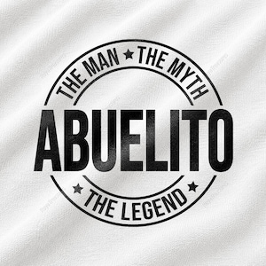 May include: A white fabric surface featuring a black circular graphic print. The design includes the text THE MAN THE MYTH in an arc at the top, the word ABUELITO in large bold letters in the center, and THE LEGEND at the bottom with stars.