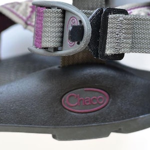 May include: Close-up of a gray and black sandal with purple accents. The sandal features a durable sole and adjustable straps. The brand name "Chaco" is visible in a purple oval on the side of the sandal.