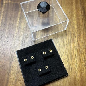 May include: A clear acrylic box with a black geometric knob on top, next to a black display stand with four pairs of gold-colored stud earrings. The earrings are set on a black velvet-like surface. The background is a wooden surface.