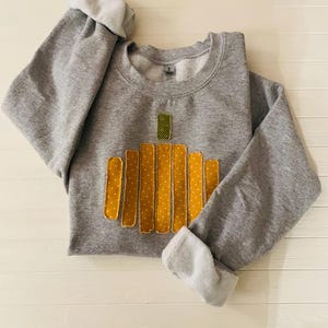 以下が含まれることがあります： グレーのトレーナーに、布の切れ端で作ったかぼちゃのデザインが施されています。かぼちゃは白のドット模様の黄色で、緑色の茎が付いています。