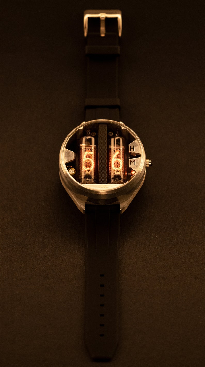 May include: A futuristic wristwatch with a black band and a silver-colored case. The watch face features two glowing orange tubes displaying the numbers "5" and "6". The watch has a buckle closure and is set against a dark background.