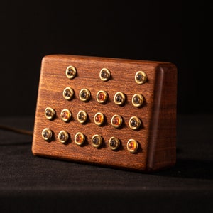 May include: A rectangular wooden object with a dark finish, featuring multiple small, round, amber-colored lights set within brass-colored bezels. The lights are arranged in a grid pattern on the front of the object. A wire extends from the side.
