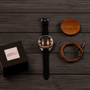 May include: A silver-cased watch with a black band displays the number 6 in orange. The watch is next to a brown wooden coaster, a brown USB cable, and a black box with a white label that reads "NEON GEAR".