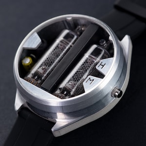 May include: A silver-colored wristwatch with a black band. The watch face is open, revealing two glass tubes with internal components. The watch has the letters "M" and "H" on the face.