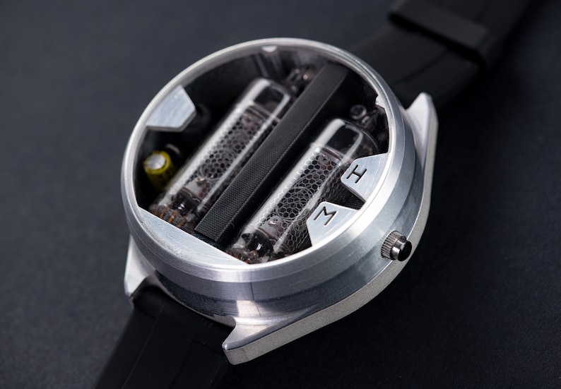 May include: A silver-colored wristwatch with a black band. The watch face is open, revealing two glass tubes with internal components. The watch has the letters "M" and "H" on the face.