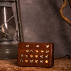 May include: A rectangular wooden block with brass-colored accents and small, illuminated orange lights. The block is on a wooden surface, with a vintage lantern and a branch in the background. The lights are arranged in a grid pattern.