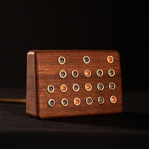 May include: A rectangular wooden block with a dark brown finish. The block features multiple small, round, gold-colored accents, some of which have orange-colored centers. A gold-colored cord extends from the side of the block.