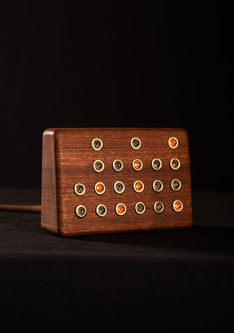 May include: A rectangular wooden block with a dark brown finish. The block features multiple small, round, gold-colored accents, some of which have orange-colored centers. A gold-colored cord extends from the side of the block.