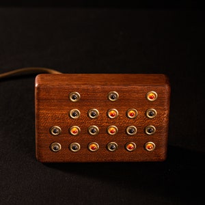 May include: A rectangular wooden box with multiple small, round buttons. The buttons are arranged in rows and columns, with some buttons having a red center and others a black center. The box has a brass-colored trim around each button.