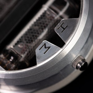 May include: Close-up of a silver-colored device with the letters "M" and "H" etched into the metal. The device has a cylindrical shape with a small knob on the side and a dark interior.