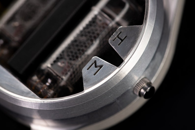 May include: Close-up of a silver-colored device with the letters "M" and "H" etched into the metal. The device has a cylindrical shape with a small knob on the side and a dark interior.