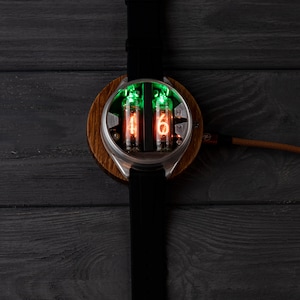 May include: A unique wristwatch featuring a round wooden base and a stainless steel case. The watch displays time with glowing orange and green numerals within glass tubes. It has a black leather strap and a silver buckle.